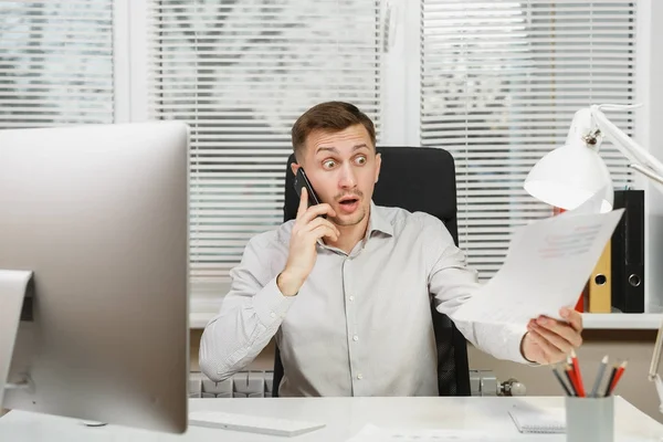 Serious angry stress business man in shirt sitting at the desk, talking, swearing and screaming on mobile phone, resolving issues, working at computer with modern monitor, documents in light office.