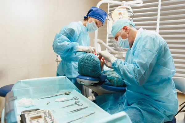 Manliga gamla professionell tandläkare kirurg i uniform och kvinnlig assistent hjälper till att utföra drift installera tandimplantat tänder av kvinnlig patient på klinik ljus kontor med moderna verktyg utrustning. — Stockfoto