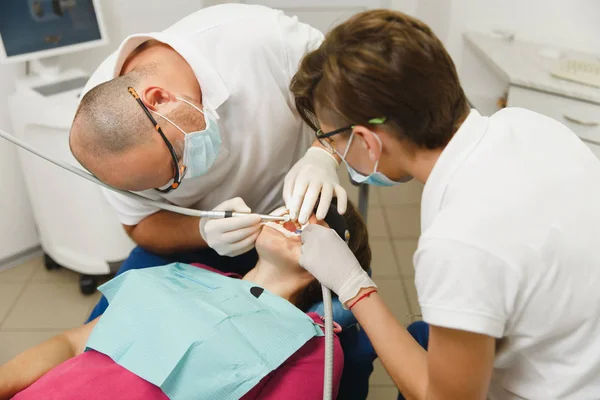 Processen att förbereda och installera keramiska tandkrona. Manliga professionella tandläkare och kvinnlig assistent hjälper till att behandla tänderna av kvinnlig patient på klinik ljus kontor med moderna verktyg utrustning. — Stockfoto