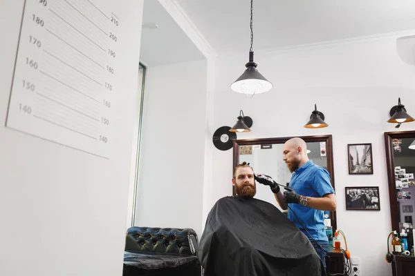 Cabeleireiro profissional masculino servindo cliente por clipper. Gengibre bonito jovem elegante brutal com barba grande grossa e cabelo curto ficando moderno corte de cabelo em capa preta. Sala de barbeiro branco claro — Fotografia de Stock