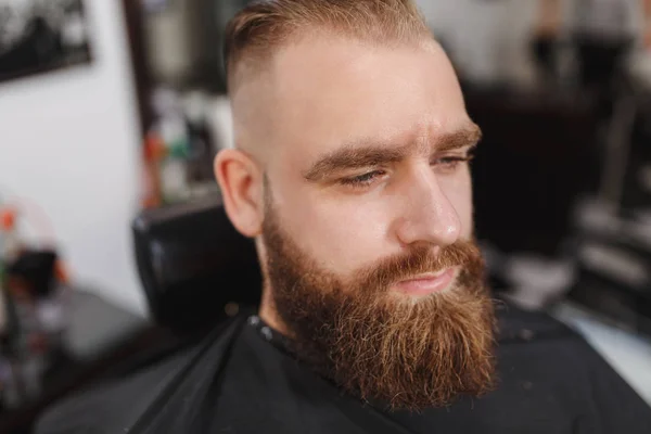 Männlicher professioneller Friseur, der Kunden bedient. Ingwer gutaussehender brutaler, stylischer junger Mann mit dickem großen Bart und kurzen Haaren, der sich eine trendige Frisur in schwarzem Umhang schneidet. heller weißer Friseursalon — Stockfoto