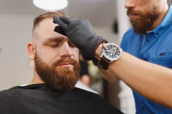 Мужчина, профессиональный парикмахер, обслуживающий клиента, стрижет брови ножницами. Рыжий стильный молодой человек с густой бородой и короткими волосами, стригущийся в черном плаще. Парикмахерская — стоковое фото