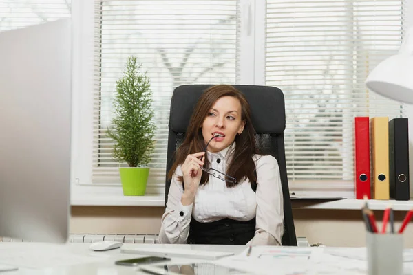 Hermosa sonriente sexy mujer de negocios de cabello castaño en traje sentado en el escritorio con tableta, teléfono móvil, lámpara blanca, trabajando en el ordenador con monitor moderno con documento en la oficina de luz, mirando a un lado — Foto de Stock