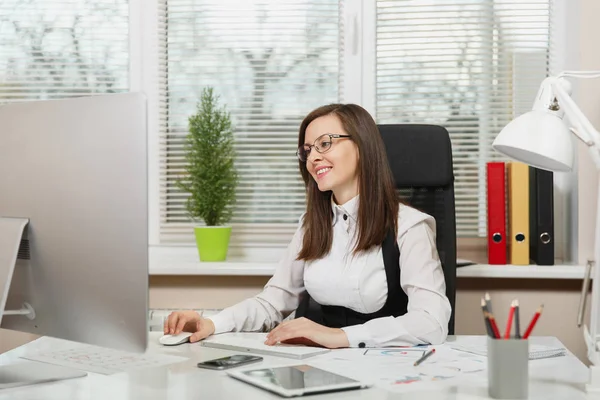 Beautiful smiling brown-hair business woman in suit and glasses sitting at the desk with tablet, working at computer with modern monitor with documents in light office, looking aside — Stock Photo, Image