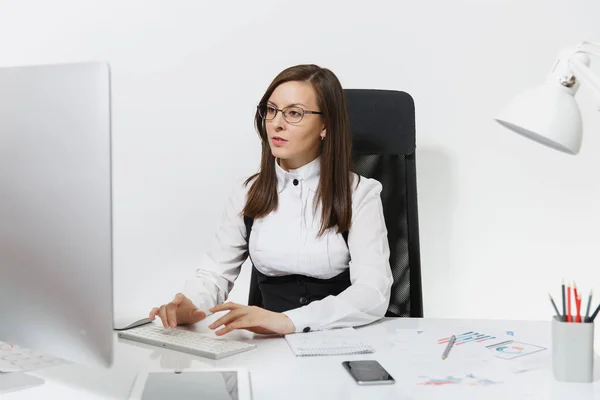 Beautiful serious and engrossed brown-hair business woman in suit and glasses sitting at the desk, working at computer with modern monitor with documents in light office, on white background — Stock Photo, Image