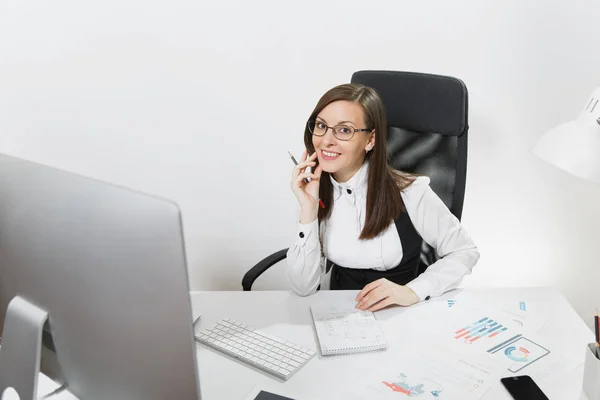 Beautiful smiling brown-hair business woman in suit and glasses sitting at the desk, working at computer with modern monitor with documents in light office, looking at the camera on white background — Stock Photo, Image