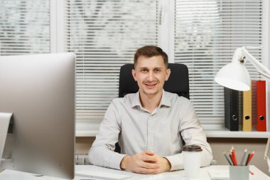 Modern monitör, lamba, klasörleri, kahve veya çay, bilgisayarda çalışan yakışıklı gülümseyen iş tişörtlü adam masada oturan pencere arka plan üzerinde ışık Office belgeleri. Yöneticisi veya alt.