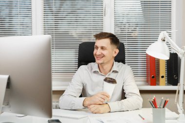 Modern İzleyicisi ile bilgisayar, belge penceresi arka plan üzerinde ışık ofiste çalışan yakışıklı gülümseyen iş tişörtlü adam fincan kahve ya da çay, ile masaya oturuyor. Yöneticisi veya alt.