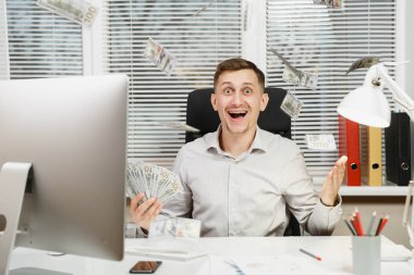 Çalışma ile modern sınıf başkanı bilgisayar, pencere arka plan üzerinde ışık office belgelerinde konusunda mutlu gülümseyen iş tişörtlü adam çok nakit para ile masaya oturuyor. Yöneticisi veya işçi kazanan.