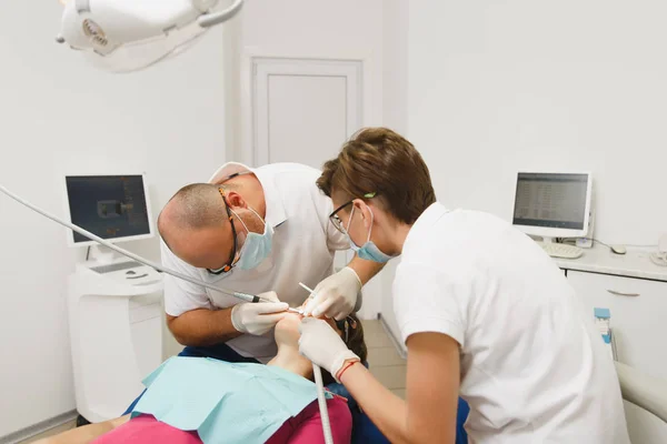 Processen att förbereda och installera keramiska tandkrona. Manliga professionella tandläkare och kvinnlig assistent hjälper till att behandla tänderna av kvinnlig patient på klinik ljus kontor med moderna verktyg utrustning. — Stockfoto