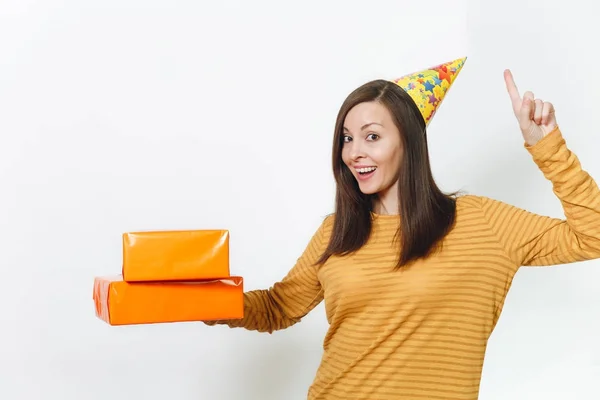 Весела молода щаслива жінка в жовтому одязі, капелюх вечірки на день народження, що тримає помаранчеві подарункові коробки з подарунком, святкуючи свято, вказуючи індекс пальця догори ногами на білому тлі ізольовані для реклами . — стокове фото