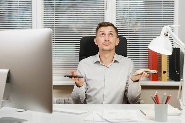 Pensativo hombre de negocios confuso en camisa sentado en el escritorio elegir entre el teléfono móvil y una gran cantidad de dinero en efectivo, trabajando en la computadora con monitor moderno, documentos en la oficina de luz. Gerente o trabajador . — Foto de Stock