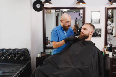 Erkek profesyonel kuaför hizmet istemci tarafından kesme makinesi. Zencefil acımasız şık yakışıklı genç kalın büyük sakal ve kısa saç Siyah pelerinli trendy saç kesimi almak. Işık beyaz Kuaför Salonu Oda