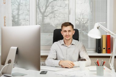 Pencere arka plan üzerinde ışık ofiste masa başında oturan gömlek ciddi ve dalmış iş adamı ile modern monitör, klasörler, kahve ya da çay, bilgisayarda çalışan makaleler. Yöneticisi veya alt.