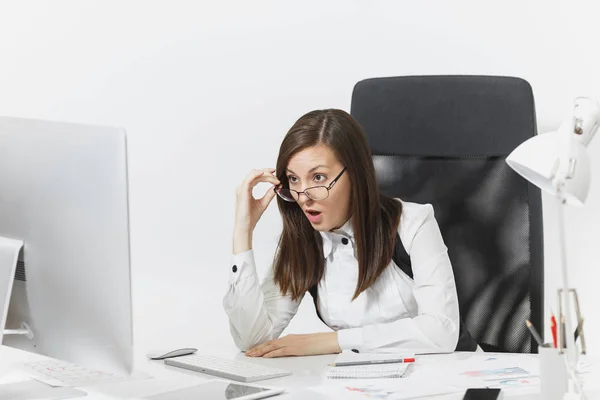 Beautiful surprised and indignant brown-hair business woman in suit and glasses sitting at the desk, working at computer, looking to modern monitor, with documents in light office, on white background — Stock Photo, Image