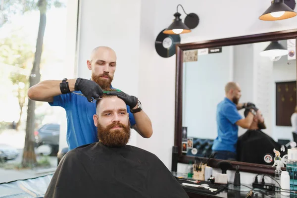 Мужчина профессиональный парикмахер, обслуживающий клиента ножницами. Рыжий красивый грубый стильный молодой человек с густой бородой, короткие волосы, модная стрижка в черном плаще. Светло-белая парикмахерская . — стоковое фото