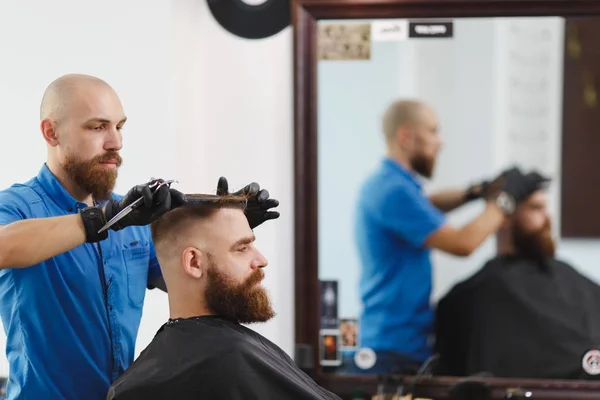 Männliche professionelle Friseur bedient Kunden mit der Schere. Ingwer gutaussehender brutaler, stylischer junger Mann mit dickem, dickem Bart, kurzen Haaren und modischem Haarschnitt in schwarzem Umhang. heller weißer Friseursalon. — Stockfoto