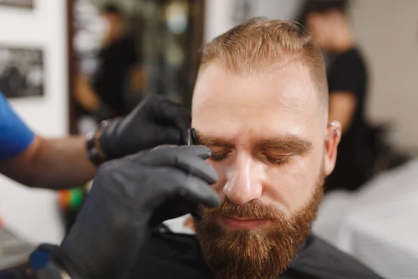 Male professional hairdresser serving client, shearing eyebrows with scissors. Ginger stylish young man with thick big beard and short hair getting trendy haircut in black cape. Light barber shop room