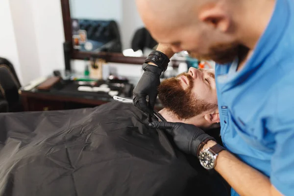 Cabeleireiro profissional masculino que serve o cliente, barbeando barba grande grossa navalha reta. Gengibre bonito jovem elegante com cabelo curto ficando moderno corte de cabelo, capa preta. Sala de barbeiro branco claro — Fotografia de Stock