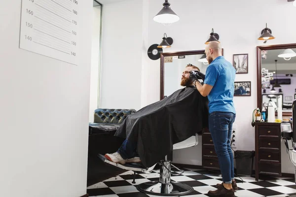 Cabeleireiro profissional masculino servindo cliente por clipper. Gengibre bonito jovem elegante brutal com barba grande grossa e cabelo curto ficando moderno corte de cabelo em capa preta. Sala de barbeiro branco claro — Fotografia de Stock