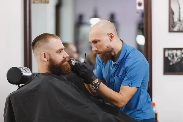 Мужчина профессиональный парикмахер, обслуживающий клиента, бреющий толстую большую бороду бритвой. Рыжий красивый стильный молодой человек с короткой стрижкой, черная накидка. Светло-белая парикмахерская — стоковое фото