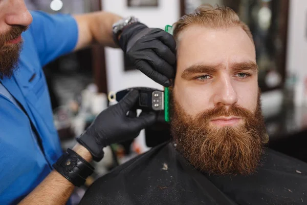 Крупный план мужского профессионального парикмахера, обслуживающего клиента с толстой большой бородой клиппером. Рыжий красивый жестокий стильный молодой человек, стригущийся в черном плаще. Светло-белая парикмахерская — стоковое фото