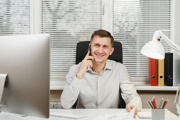 Homem de negócios sorridente na camisa que se senta na mesa, falando no telefone celular, conduzindo a conversação agradável, trabalhando no computador com o monitor moderno, documentos no escritório leve no contexto de janela . — Fotografia de Stock