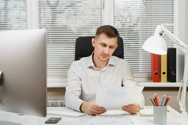 Serious and engrossed business man in shirt sitting at the desk, working at computer with modern monitor, folders, coffee or tea, documents in light office on window background. Manager or worker.