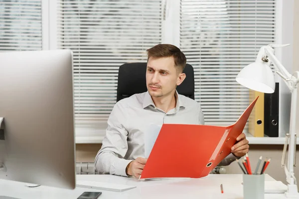 Beau homme d'affaires sérieux et absorbé en chemise assis au bureau, travaillant à l'ordinateur avec moniteur moderne, dossier rouge, documents dans le bureau léger sur fond de fenêtre. Directeur ou travailleur . — Photo