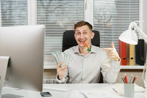 Beau homme d'affaires amusant en chemise assis au bureau avec beaucoup d'argent comptant, tuyau d'anniversaire, travail à l'ordinateur avec moniteur moderne, documents dans le bureau léger sur fond de fenêtre. Directeur ou travailleur — Photo