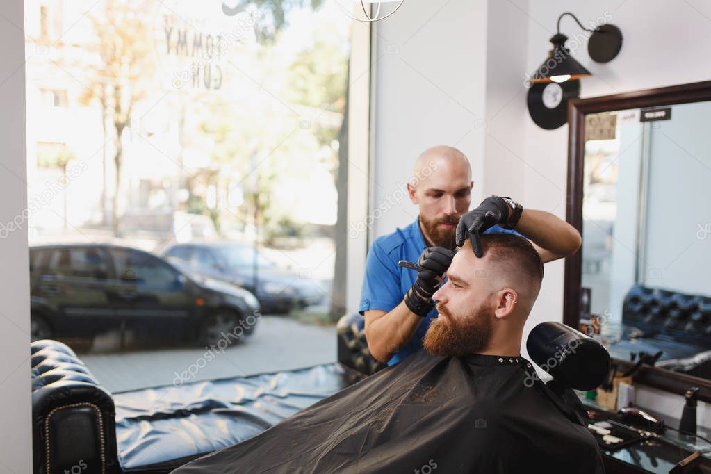 Male professional hairdresser serving client, shaving thick big beard straight razor. Ginger handsome stylish young man with short hair getting trendy haircut, black cape. Light white barber shop room
