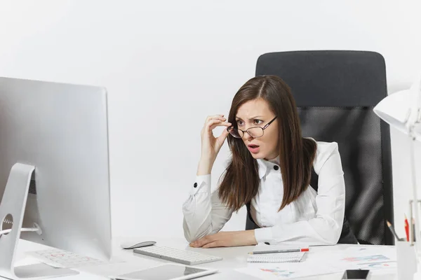 Beautiful surprised and indignant brown-hair business woman in suit and glasses sitting at the desk, working at computer, looking to modern monitor, with documents in light office, on white background — Stock Photo, Image