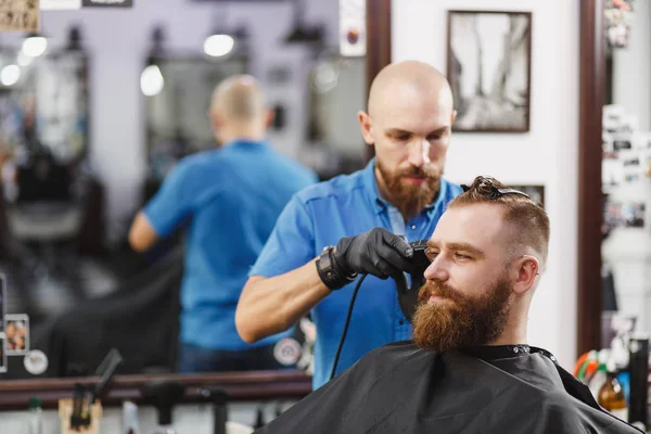Male professional hairdresser serving client by clipper. Ginger handsome brutal stylish young man with thick big beard and short hair getting trendy haircut in black cape. Light white barber shop room — Stock Photo, Image