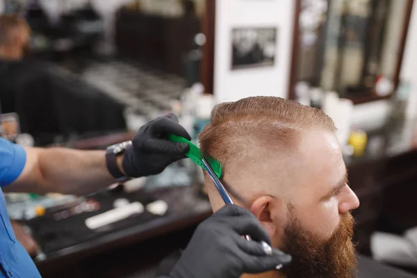 Close up de cabeleireiro profissional masculino servindo cliente por tesoura. Gengibre bonito jovem elegante com barba grande, cabelo curto ficando moderno corte de cabelo em capa preta. Sala de barbeiro branco claro . — Fotografia de Stock