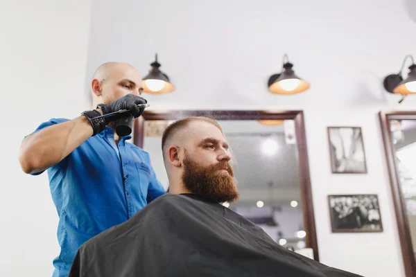 Stock image Male professional hairdresser serving client by scissors. Ginger handsome brutal stylish young man with thick big beard, short hair getting trendy haircut in black cape. Light white barber shop room.