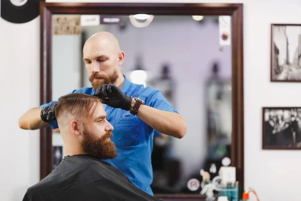 Male professional hairdresser serving client by scissors. Ginger handsome brutal stylish young man with thick big beard, short hair getting trendy haircut in black cape. Light white barber shop room. — Stock Photo, Image