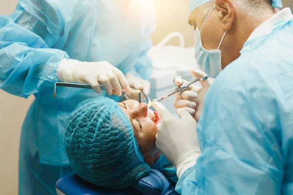 Male old professional dentist surgeon in uniform and female assistant helps to performing operation install dental implant teeth of woman patient in clinic light office with modern tools equipment.