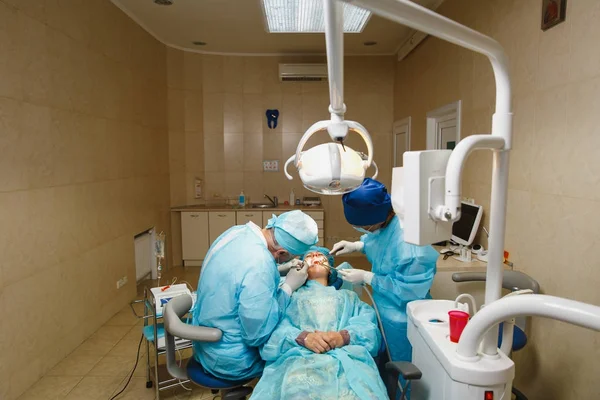 Male old professional dentist surgeon in uniform and female assistant helps to performing operation install dental implant teeth of woman patient in clinic light office with modern tools equipment.