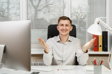 Eller, bilgisayarda çalışan modern monitör, lamba, klasörler, belge penceresi arka plan üzerinde ışık Office ile yayılan yakışıklı gülümseyen iş tişörtlü adam masada oturuyor. Yöneticisi veya alt.