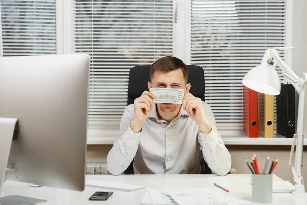 Hombre de negocios guapo y serio en camisa sentado en el escritorio escondido detrás de dinero en efectivo, trabajando en la computadora con monitor moderno, documentos en la oficina de luz en el fondo de la ventana. Gerente o trabajador . —  Fotos de Stock