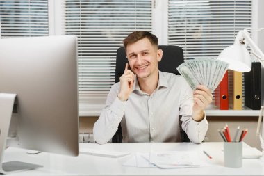 Bilgisayarda çalışan modern monitör, lamba, ışık Office belgeleri ile yakışıklı gülümseyen iş tişörtlü adam çok nakit para, cep telefonuyla konuşurken masasında oturuyor. Yöneticisi veya alt.