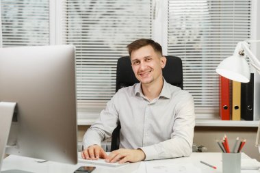 Bilgisayar başında modern monitör ile çalışan yakışıklı gülümseyen iş tişörtlü adam masada oturan eller klavye, klasörler, belge penceresi arka plan üzerinde ışık ofiste. Yöneticisi veya alt.