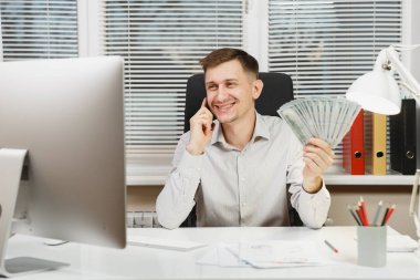Bilgisayarda çalışan modern monitör, lamba, ışık Office belgeleri ile yakışıklı gülümseyen iş tişörtlü adam çok nakit para, cep telefonuyla konuşurken masasında oturuyor. Yöneticisi veya alt.