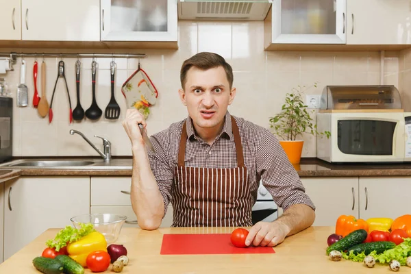 Angry crazy caucasian young man in apron, brown shirt sitting at