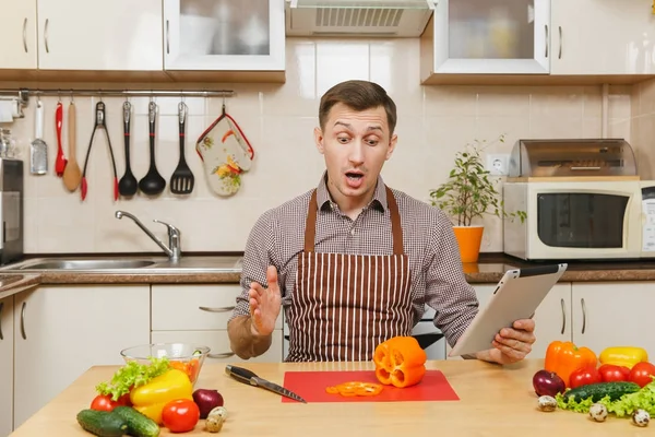 Όμορφος Καυκάσιος νεαρός άνδρας σε μια ποδιά, καφέ πουκάμισο που κάθεται στο τραπέζι, ψάχνει συνταγή σε δισκίο, cuting λαχανικό για σαλάτα με μαχαίρι σε ελαφριά κουζίνα. Δίαιτα έννοια. Το μαγείρεμα στο σπίτι. Προετοιμασία φαγητού — Φωτογραφία Αρχείου