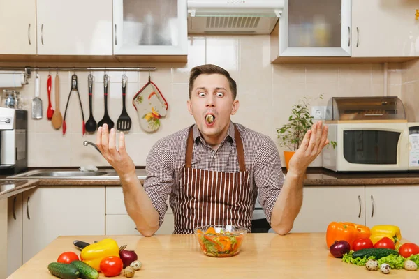 Kase ve Bıldırcın yumurta içinde ağız içinde sebze salatası ile masada oturmuş, hafif mutfak elinde yayılan önlük, kahverengi gömlekli eğlenceli beyaz genç adam. Diyet kavramı. Evde yemek. Yemek hazırlamak.