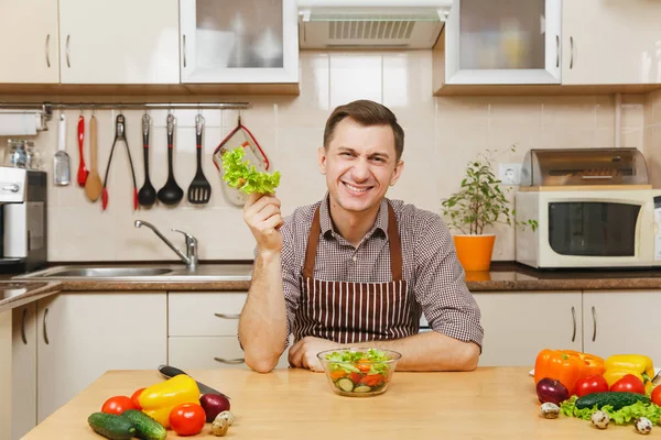 Schöner kaukasischer junger Mann in Schürze, braunes Hemd sitzt am Tisch und schneidet und zerreißt Salatgemüse für Salat mit Messer in heller Küche. Diätkonzept. Kochen zu Hause. Essen zubereiten. — Stockfoto