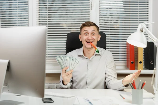 Bello uomo d'affari divertente in camicia seduto alla scrivania con un sacco di soldi in contanti, tubo di compleanno, lavorare al computer con monitor moderno, documenti in ufficio luce sullo sfondo della finestra. Responsabile o lavoratore — Foto Stock