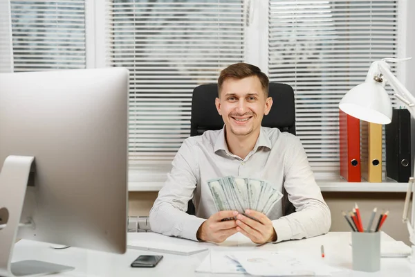 Feliz hombre de negocios sonriente en camisa sentado en el escritorio con mucho dinero en efectivo, trabajando en la computadora con monitor moderno, documentos en la oficina de luz en el fondo de la ventana. Gerente o trabajador ganador . —  Fotos de Stock