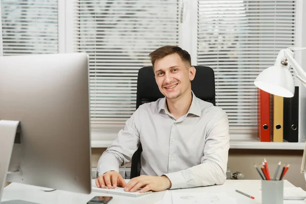 Красивий усміхнений бізнесмен у сорочці, що сидить за столом, працює за комп'ютером з сучасним монітором, руки на клавіатурі, папки, документи у світлому офісі на фоні вікна. Менеджер або працівник . — стокове фото